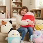 Happy little girl sitting on stairs, hugging a large strawberry Squishmallows plush, surrounded by a collection of other assorted Squishmallows characters like a banana and cat for a toy review.
